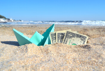 Blue small paper boat and three dollar paper bills half in sand on beach near sea on summer sunny...