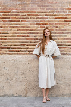 Full Length Of Dreamy Redhead Woman In Dress Posing Near Brick Wall On European Street.