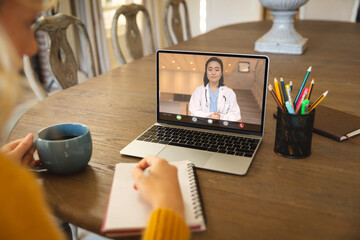 Asian female doctor on video call talking to caucasian businesswoman writing on book at desk - obrazy, fototapety, plakaty