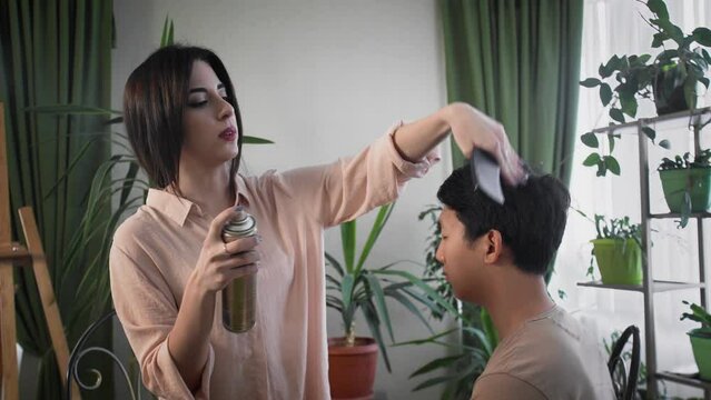 The stylist creates an image of an Asian guy for a photo shoot. A young woman fixes her boyfriend's hair with hairspray.
