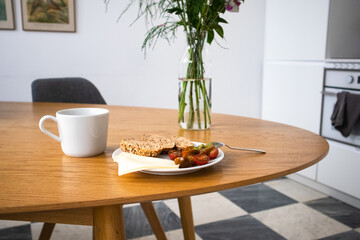 Cozy home kitchen. A plate of cherry tomatoes, sliced bread, and a white ceramic mug stands on a wooden oval table next to a bouquet of flowers against a white wall. Eating indoors. Tasty healthy food