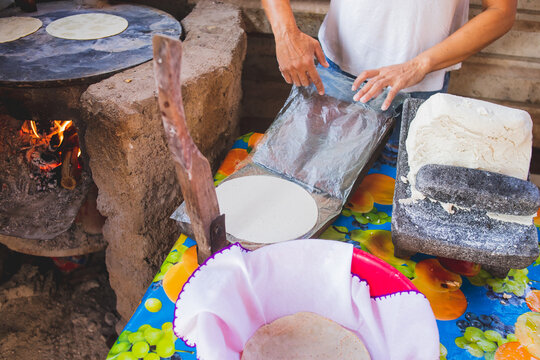 Mujer mexicana torteando maza de ma&iacute;z en un metate y una estufa de le&ntilde;a para hacer tortillas caseras	

