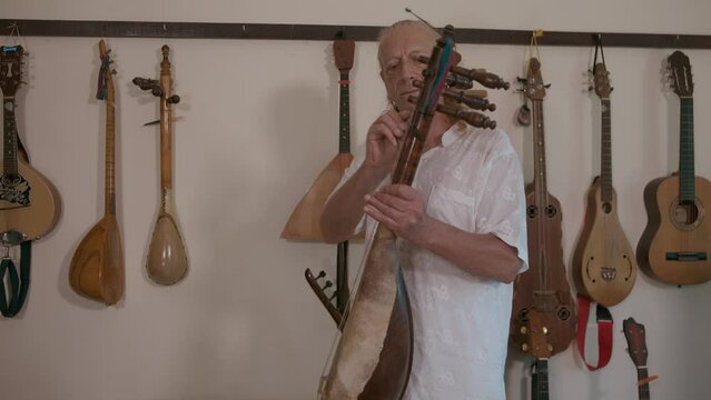 Elderly caucasian man playing african stringed instrument Guembri in his home studio. Behind him on the wall is a collection of oriental stringed musical instruments.