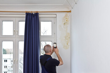 Man taking a photo of a big patch of mold or mildew growing behind the drapes of a white external wall in an old house. - obrazy, fototapety, plakaty