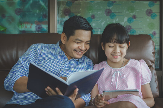 Father and daughter are reading a book and using tablet while sitting on sofa at home.Concept of Father's day