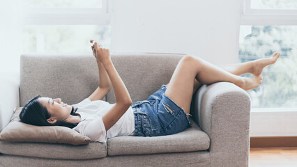 Asian woman sitting with smart phone on sofa in living room, Looking at tablet screen, Relaxing at home, Working through the Internet communication system, Happy working lifestyle.