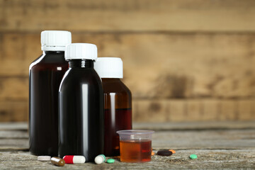 Bottles of cough syrup, measuring cup and pills on wooden table. Space for text
