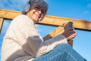 Senior adult smiling woman sitting in outdoor wearing headphones listening to music. Attractive elderly woman with denim pants relaxing enjoying free time and coffee break