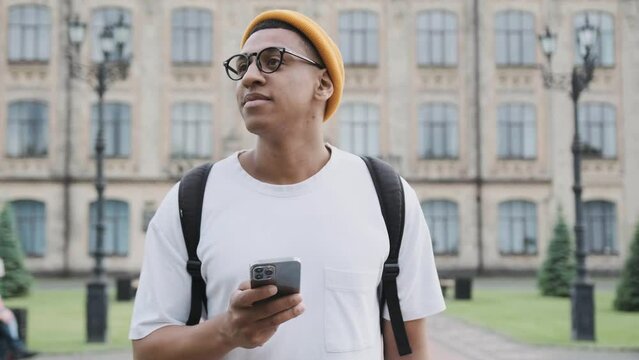 Motion scene of a young stylish African American international student walking in the campus texting on the phone