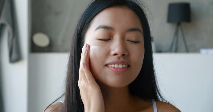 Close-up of attractive Asian woman with healthy skin, beautiful long hair, applying daily moisturizer to her face, performing facial massage to smooth during her daily morning routine in the bathroom.