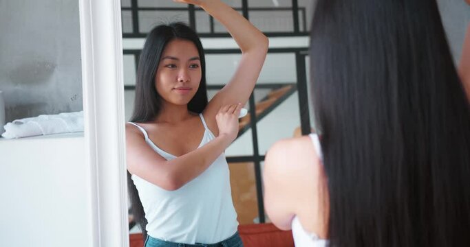 Beautiful Asian woman feeling enjoyment and happiness, smiles looking at her mirror reflection at home, applying roll-on antiperspirant deodorant on armpits. Daily hygiene routine. Body care concept
