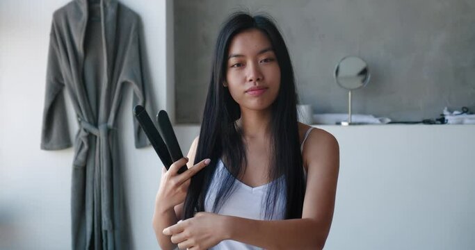 Hairstyling, hairdressing concept. Cute smiling girl straightening healthy brunette hair with flat iron. Beautiful young Asian woman with bright healthy long straight hair using hair straightener.
