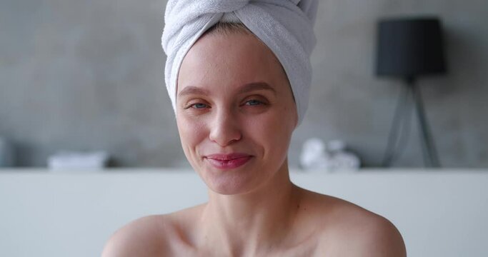 Close-up of a cheerful gorgeous blonde woman with head wrapped in white terry towel laughs and smiles with beautiful toothy smile, posing in front of the camera, standing half naked in home bathroom