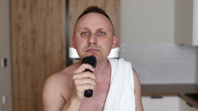 young unshaven man with towel on shoulders shaving with modern electric razor trimer while looking at camera, indoors. Daily routine male self-care. man shaves with an electric razor in the kitchen.