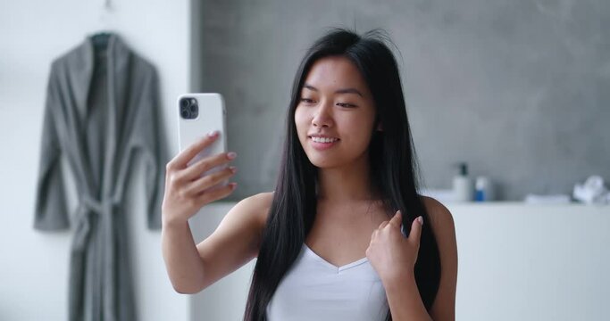 Attractive young asian woman making selfie photos using smartphone while standing in bathtub at home. Millennial girl takes pictures of herself with mobile phone in the bathroom for a beauty blog