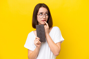 Young Ukrainian woman isolated on yellow background taking a chocolate tablet and having doubts