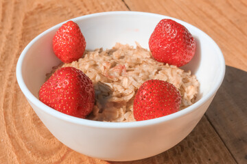 Oatmeal with strawberries in a white plate isolated on a wooden table, dissolves. Healthy food for every morning.