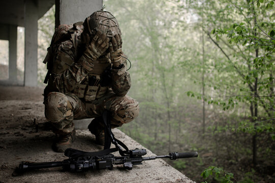 Military soldier in uniform sitting on his knees, releasing his head in front of the machine gun. Loss of a combat partner. Defeat in battle