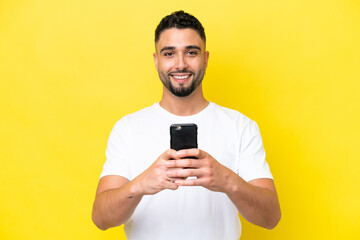 Young Arab handsome man isolated on yellow background looking at the camera and smiling while using the mobile