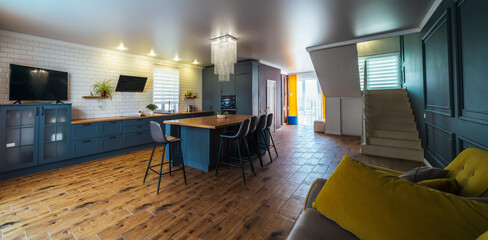 Interior of modern kitchen in blue with yellow accents and wooden floor with large table in middle of room, cooking area and sofa for relaxing.