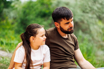 Portrait of satisfied family sitting on picnic in park forest around trees bushes. Happy middle-aged man father embracing little girl daughter, having fun. Love, family, summer, togetherness, care.