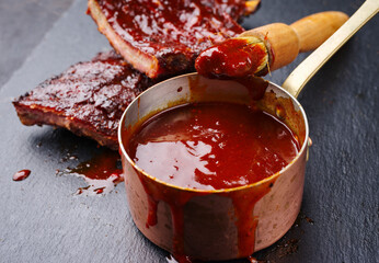 Hot and spicy barbecue sauce in a casserole as close-up with spare ribs in background - obrazy, fototapety, plakaty