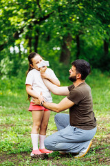 Side view of middle-aged bearded attentive man father squatting on hams, wiping daughters nose with white handkerchief during walking in summer green forest. Family, love, care, sneezing. Vertical.