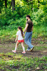 Naklejka na ściany i meble Back view of family walking in park forest around green trees, talking, having fun. Little cheerful daughter holding hand of middle-aged bearded man father. Summer activities, travelling. Vertical.