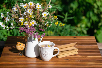Garden composition bouquet of meadow flowers, herbal tea cup, books on table in summer. Rest in garden, reading books, breakfast, rest in nature concept. Summertime in garden on backyard