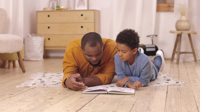 Loving african american dad with schoolboy son lying on floor and flipping cognitive encyclopedia pages at bright home. Responsible father taking part in childish educational process.