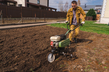 Farmer cultivates the ground soil in the garden using a motor cultivator or tiller tractor. Modern farming and technology agriculture.  - obrazy, fototapety, plakaty