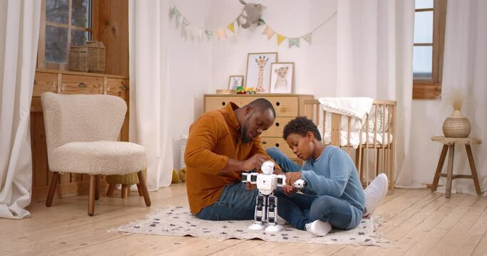 African american dad teaching little son using screwdriver for adjustment of interactive robot toy. Bearded father sharing experience and giving advices for boy while sitting on living room floor.