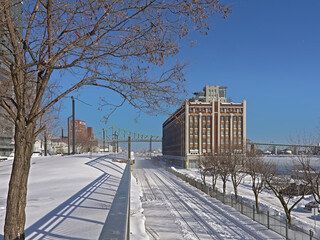 Naklejka premium Old industrial building and Jaques Cartier Bridge over Frozen Saint Lawrence river with snow on a sunny winter day in the old port of Montreal 