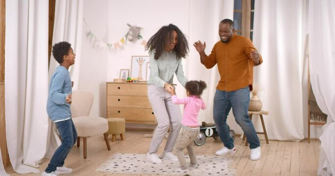 Excited Young Parents Dancing To Music With Playful Little Children At Home. Overjoyed African American Family Of Four Spending Active Free Time Together In Modern Playroom.