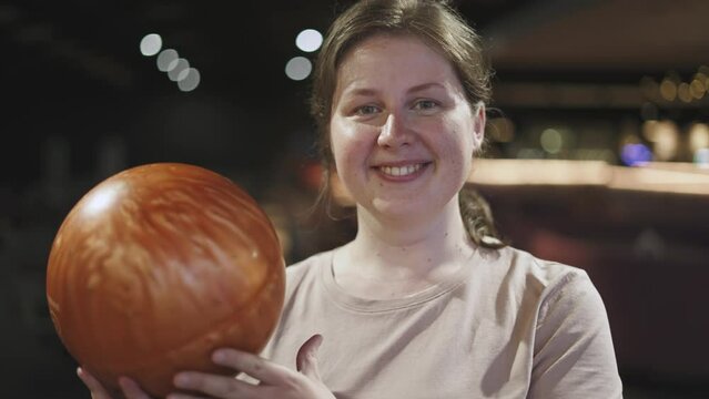Portrait of charming, positive young woman with freckles and moles on her skin in bowling alley. Young adult holding orange ball, looking at sports equipment, smiling at the camera, close-up.