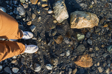 ángulo en primera persona, pantalones cafés y zapatillas deportivas blancas a la orilla del mar de aguas cristalinas con rocas y arena 