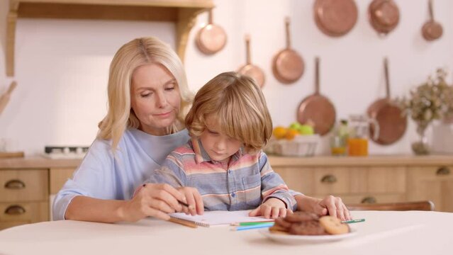 Pretty loving woman holding small attentive son on knees and helping him to draw beautiful picture in album. Happy mother with son sitting at kitchen table and using colorful pencils for painting.