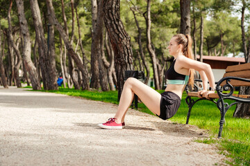 Obraz na płótnie Canvas Young caucasian woman wearing black sports bra standing on city park, outdoors concentrating on her workout, leaning on a park bench, doing triceps exercise.