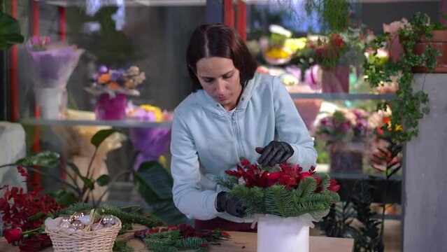 Skilled absorbed Caucasian florist inserting red leaves in bouquet base standing at table in workshop. Portrait of concentrated confident woman creating composition in flower shop indoors