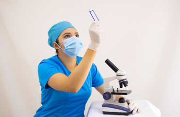 beautiful woman lab technician wearing uniform and blue surgical cap, surgical protection, analyzing samples with the microscope, on white background