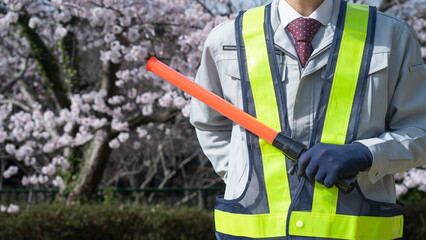 桜の背景で作業服で誘導棒を持つ人｜建設業・警備業
