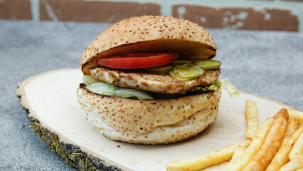 Close-up of tasty chicken burger with french fries, lettuce, tomato and cucumber pickle on a natural wooden tray front of vintage brick wall	