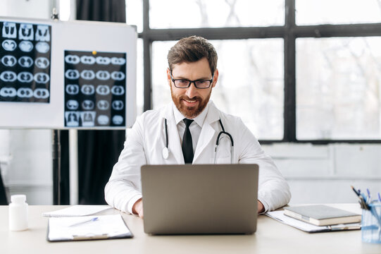 Smart proud positive caucasian male doctor of general medicine, dressed in a medical uniform and with glasses, sits at his workplace in a medical office, looking in a laptop, consults patients online