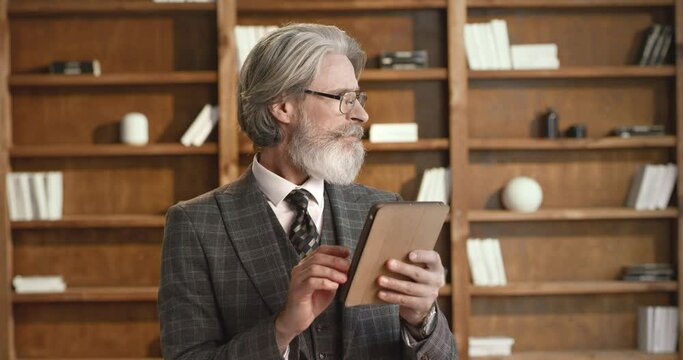 Bearded university professor in trendy glasses and formal suit working on digital tablet at modern library. Senior man checking online required list of training manuals.