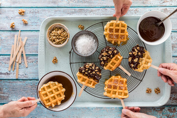 Hands holding homemade waffle pops with toppings all around. - obrazy, fototapety, plakaty