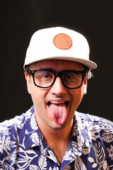 Young man looking towards the camera sticking out his tongue, wearing  glasses, white cap and blue and white Hawaiian shirt on black background.