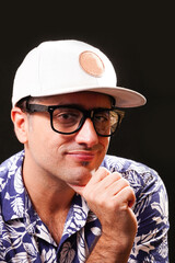 Young man looking towards the camera smiling, with glasses, white cap and blue and white Hawaiian shirt on black background.