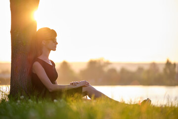 Lonely woman sitting alone on green grass lawn leaning to tree trunk on lake shore on warm evening....