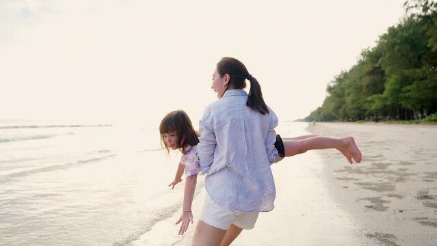 4K Happy Asian family on beach vacation. Mother carrying and playing with little daughter while walking on the beach at summer sunset. Mom and child girl kid enjoy and fun outdoor lifestyle at the sea
