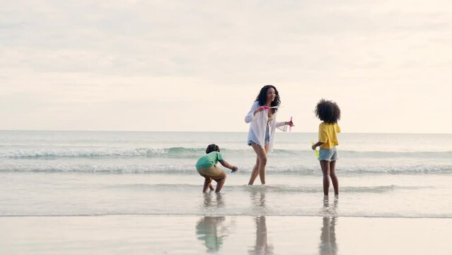 Happy African family mother with little daughter and son running and playing sea water together on the beach at summer sunset. Mom with child boy and girl enjoy outdoor lifestyle vacation at the sea